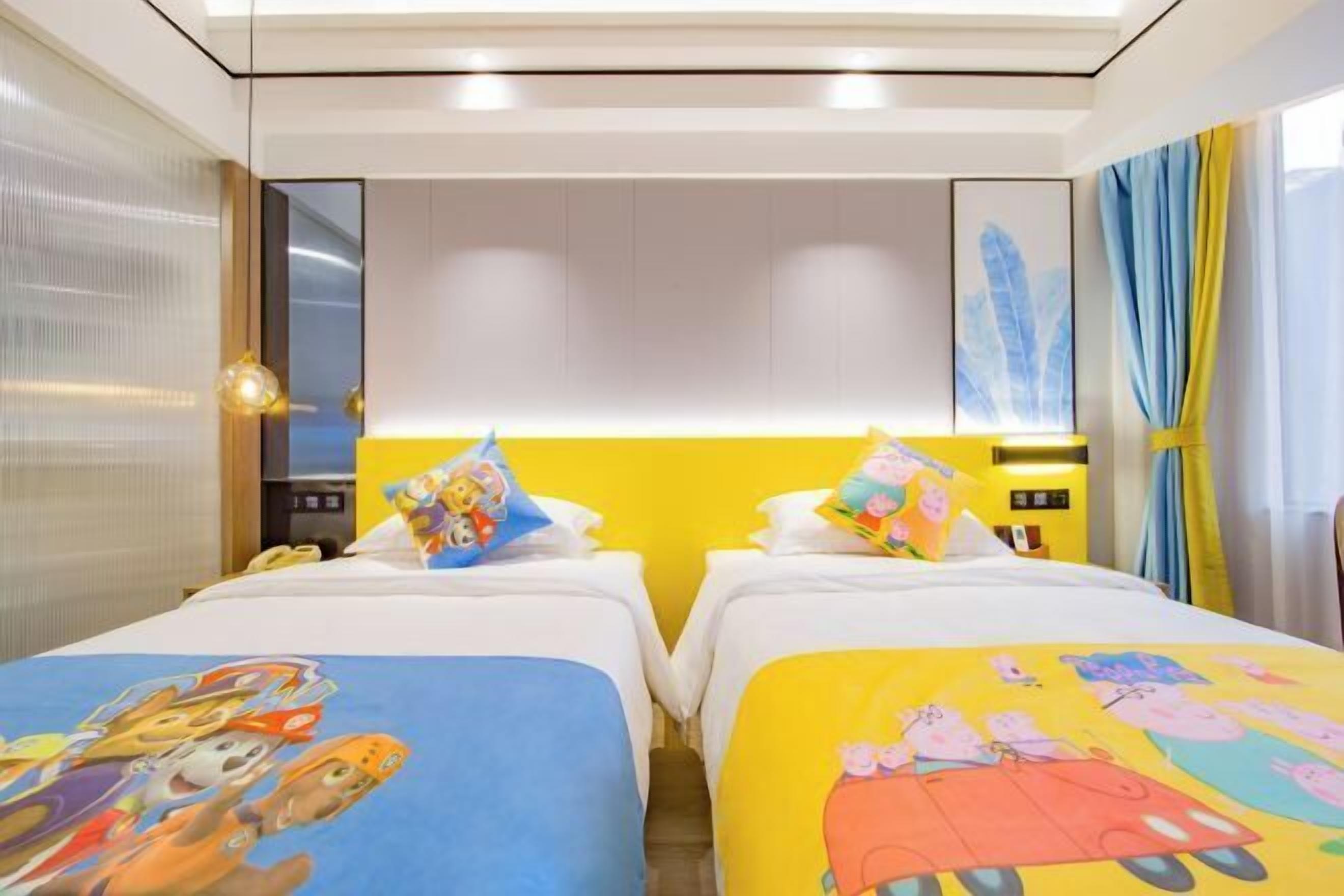 A hotel room with two beds, each with colorful bedding featuring cartoon characters. There is a bedside table with a lamp, and the room has a modern design with a neutral color scheme.