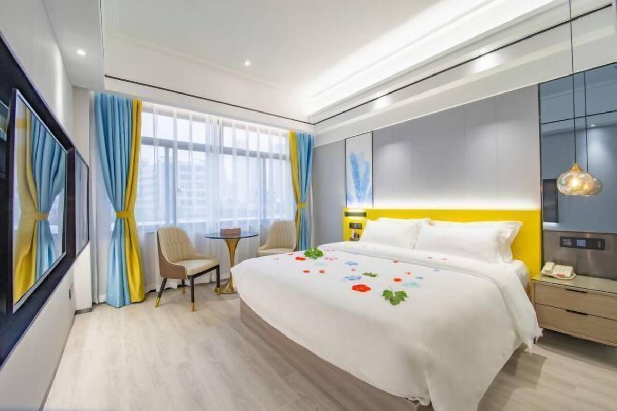 A modern hotel room with a large bed, a small table, a chair, and a bedside table with a lamp.