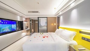 A modern hotel room with a large flat-screen TV, a bed with white bedding, a bedside table with a remote control, and a glass nightstand.
