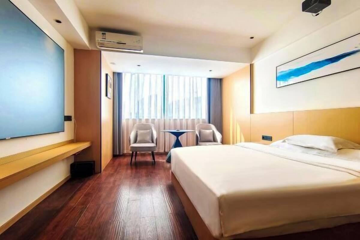 Ein Hotelzimmer mit Bett, Schreibtisch, zwei Stühlen, einem kleinen Tisch und einem großen Fenster mit Vorhängen.