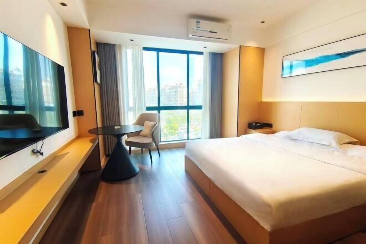 Ein Hotelzimmer mit einem großen Bett, einem Fernseher, einem kleinen Tisch und einem Sessel. Es gibt ein Fenster mit Vorhängen und ein Bild an der Wand.