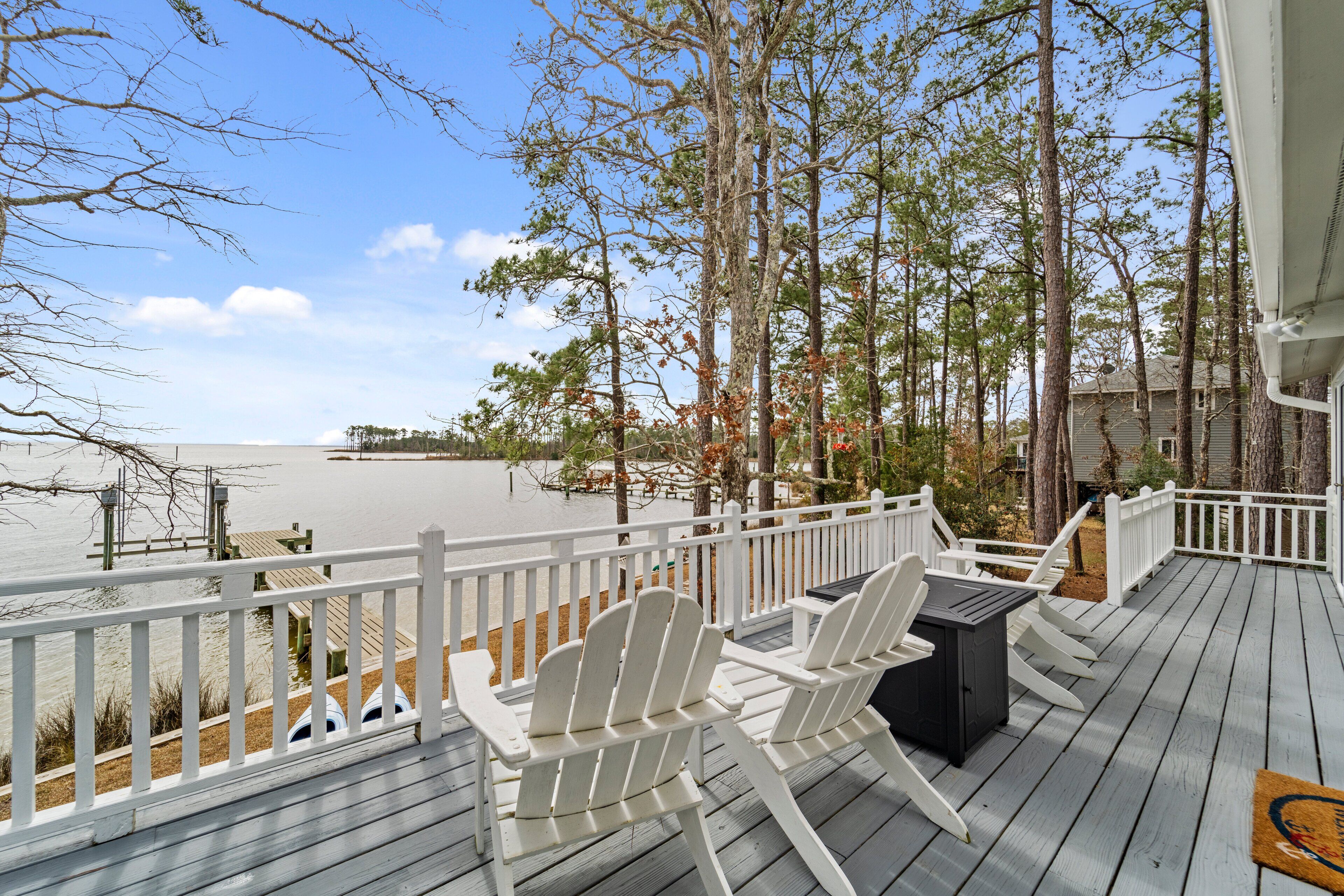 A wooden deck with white Adirondack chairs and a grill, overlooking a body of water and trees.