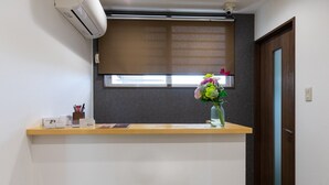 A reception desk with a vase of flowers, a window with blinds, and a door. - Mr.KINJO Happiness Resort 1 (Miyakojima)