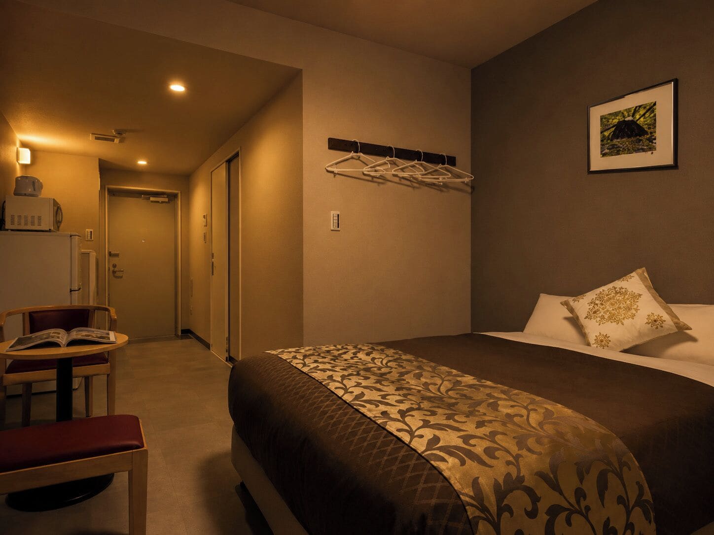 A hotel room with a bed, a chair, a small table with a book, a wall-mounted coat rack, and a framed picture on the wall.