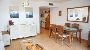 Un coin repas avec des chaises en osier et une table en bois, un meuble blanc avec des étagères ouvertes, et un jeu d’échecs posé sur le sol.