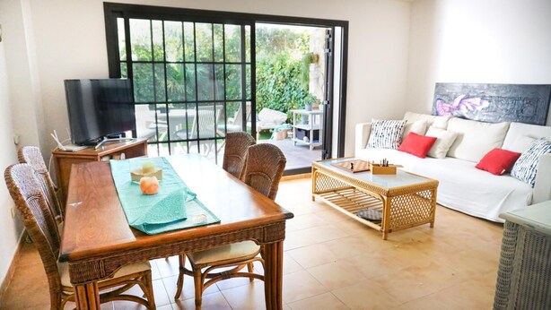 A spacious living area with a dining table, wicker chairs, a sofa, and a television. - VACAY L'Escala – Gala Duplex with Pool and Parking near Riells Beach (l'Escala)
