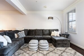 Una sala de estar con un sofá seccional, otomanas, una lámpara de pie y una ventana con persianas.
