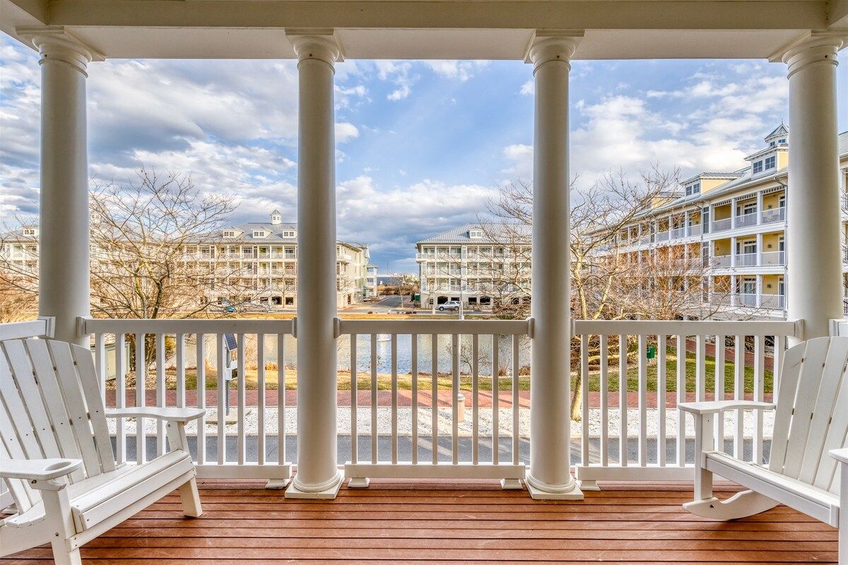 A covered porch with white columns and benches, overlooking a street with buildings and a body of water.