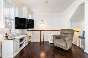A living room with a flat-screen TV, a comfortable armchair, a wooden staircase, and a white console table. - Sunset Island 13 Fountain Drive West (Ocean City)