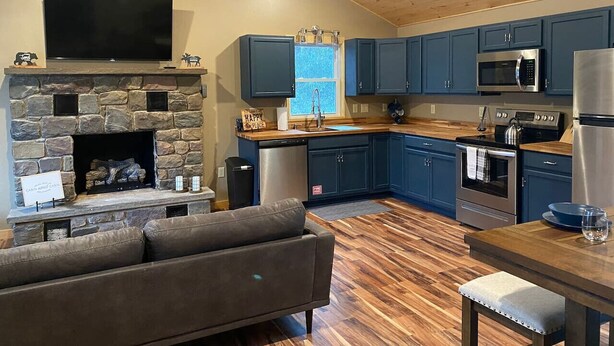 A modern kitchen with a stone fireplace, a flat-screen TV, and a dining area with a wooden table and chairs. - Mtn Rd Cottage -HOT TUB-Close 2 Camelback/Shopping (Tannersville)