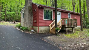 A small cabin with a chimney, a deck, and a paved driveway surrounded by trees. - Mtn Rd Cottage -HOT TUB-Close 2 Camelback/Shopping (Tannersville)
