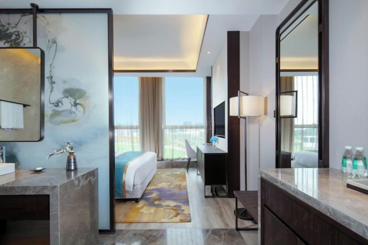 Una habitación de hotel moderna con una cama grande, un escritorio, una silla, un baño con espejo y vistas a la ciudad.