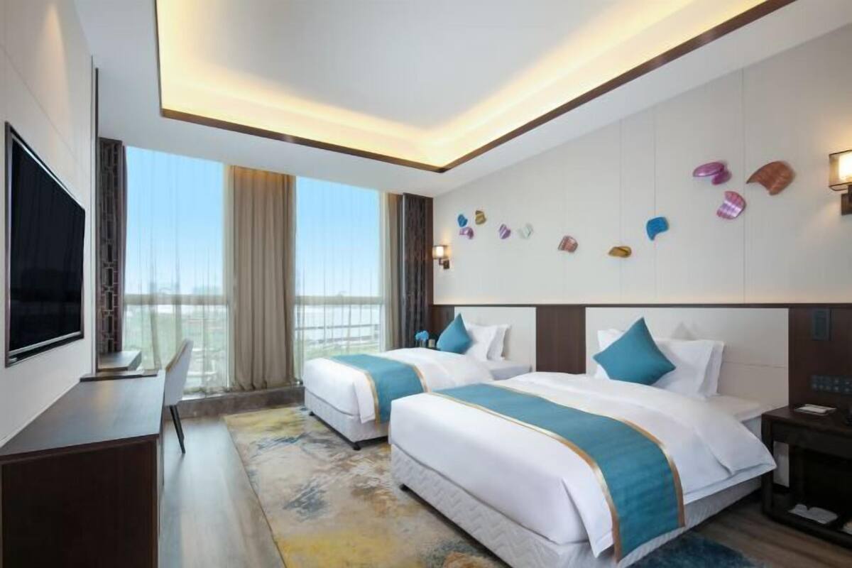 Habitació d'hotel amb dos llits, una televisió, un escriptori i un finestra gran amb cortines.