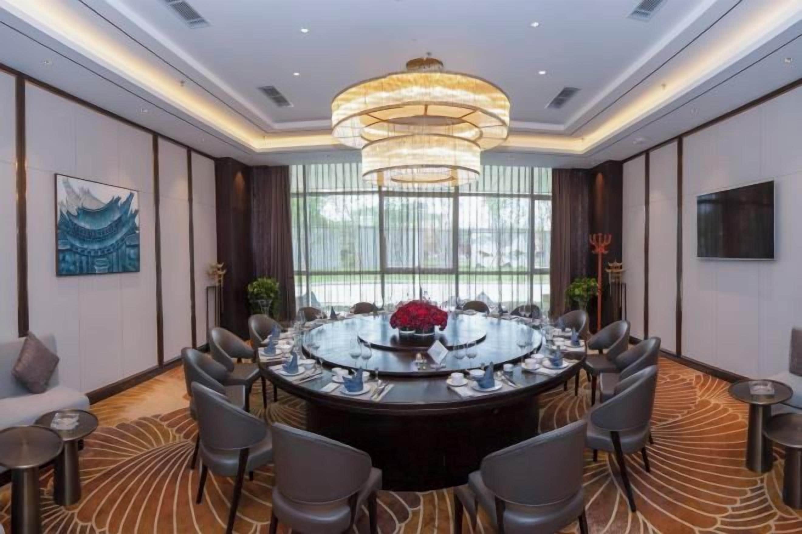 Uma sala de conferências com uma mesa redonda, cadeiras, um lustre e uma grande janela.