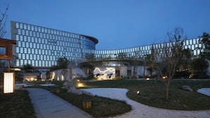 Un hotel moderno con fachada de vidrio curvo, un jardín ajardinado con senderos y una entrada destacada con obra de arte iluminada.