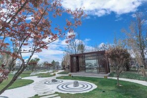 Un edificio moderno con fachada de vidrio, rodeado de un jardín ajardinado con diseños circulares de piedra y árboles.