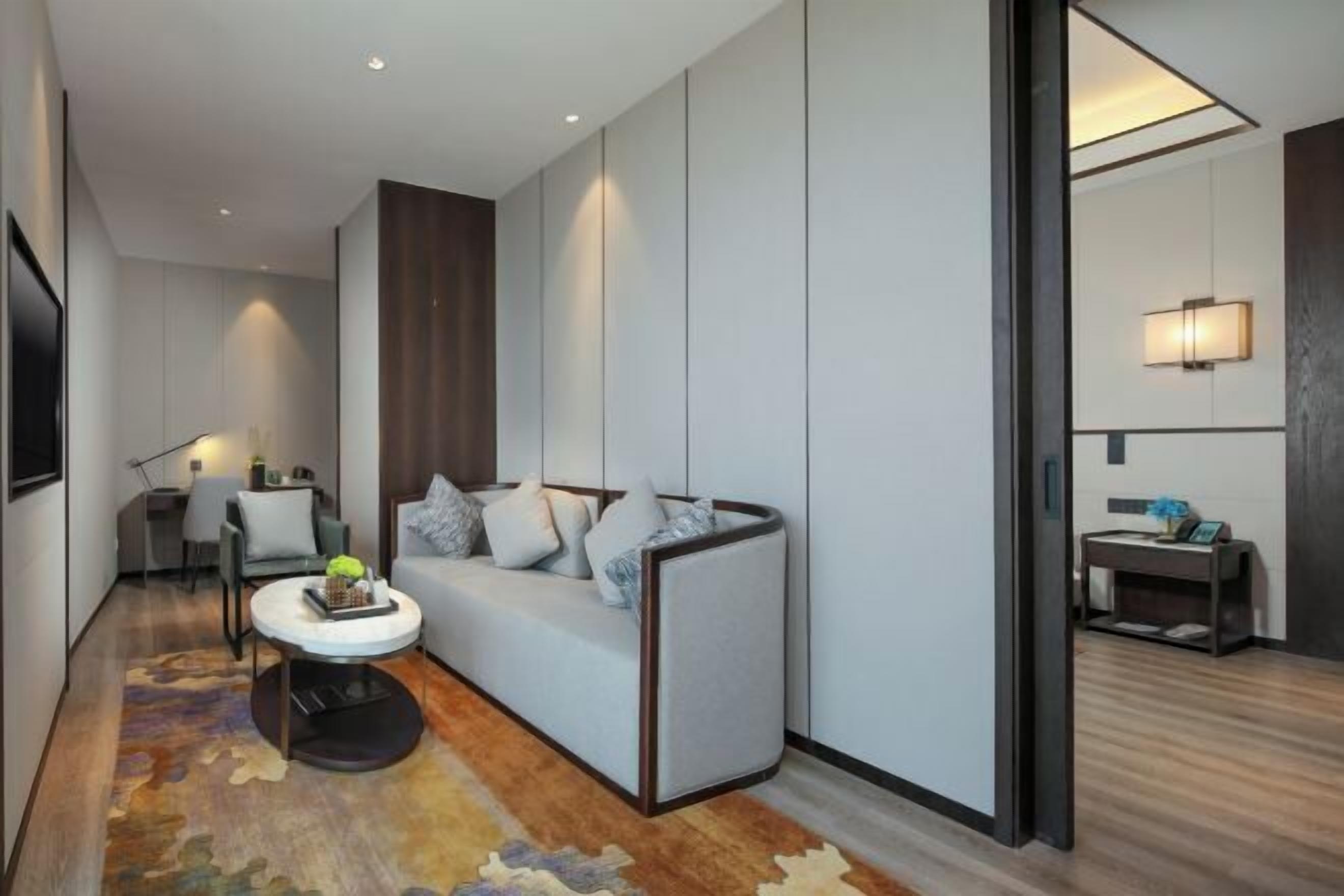 A modern hotel room with a sofa, a small round table with a decorative item, a desk with a chair, and a wall-mounted TV.