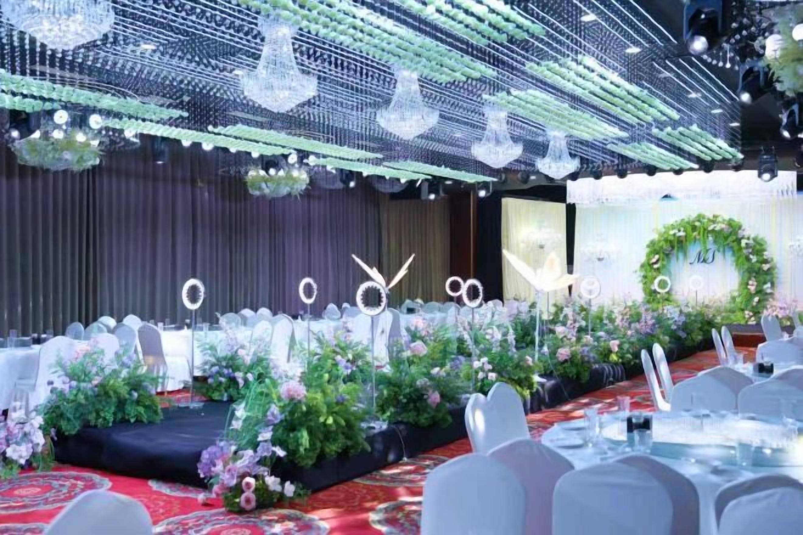 Une salle de banquet avec des tables dressées pour un événement formel, une scène agrémentée de compositions florales et un plafond décoratif.