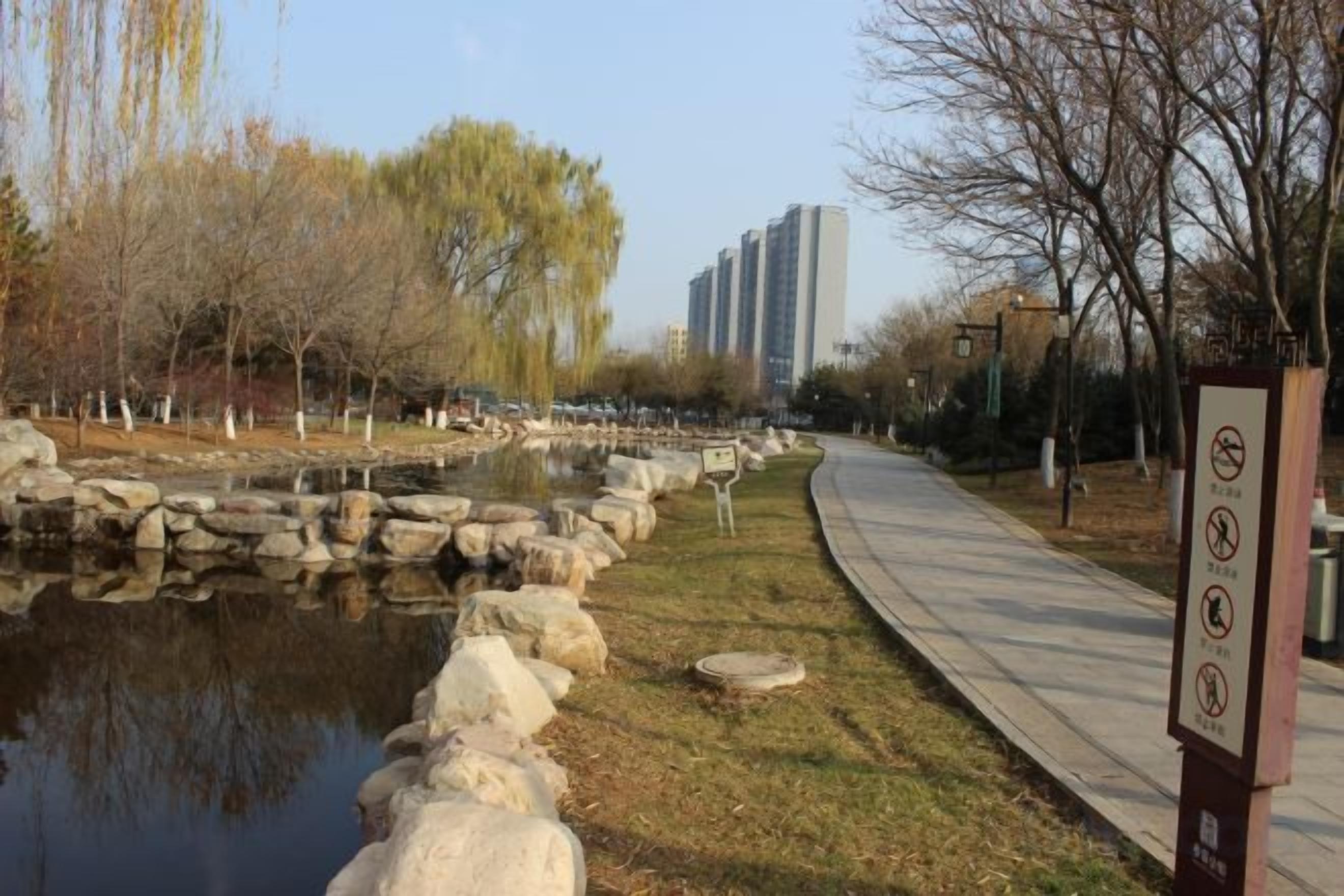 A pathway alongside a body of water with rocks, trees, and a signboard.