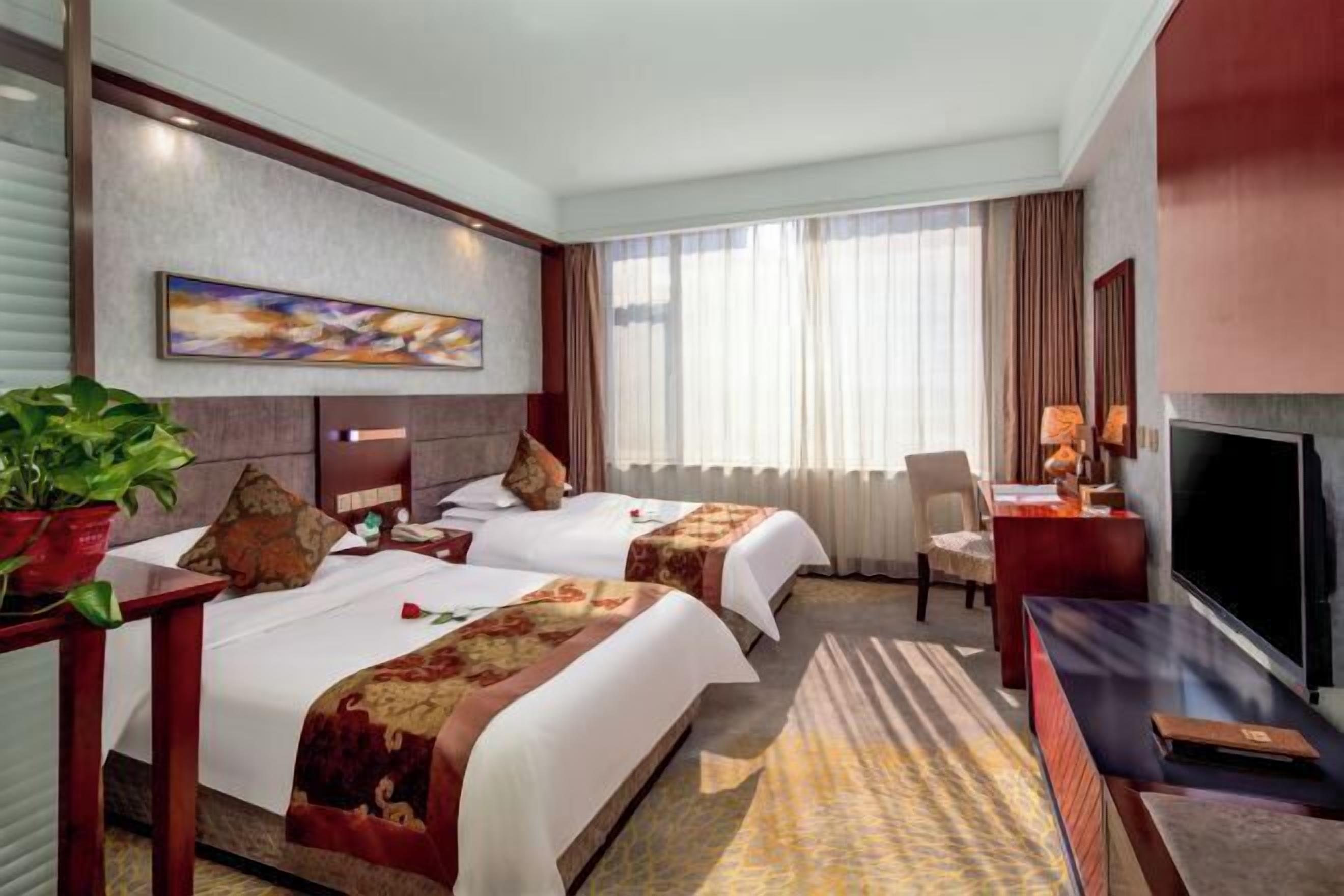 A hotel room with two beds, a TV, a desk, a chair, and a large window with curtains.