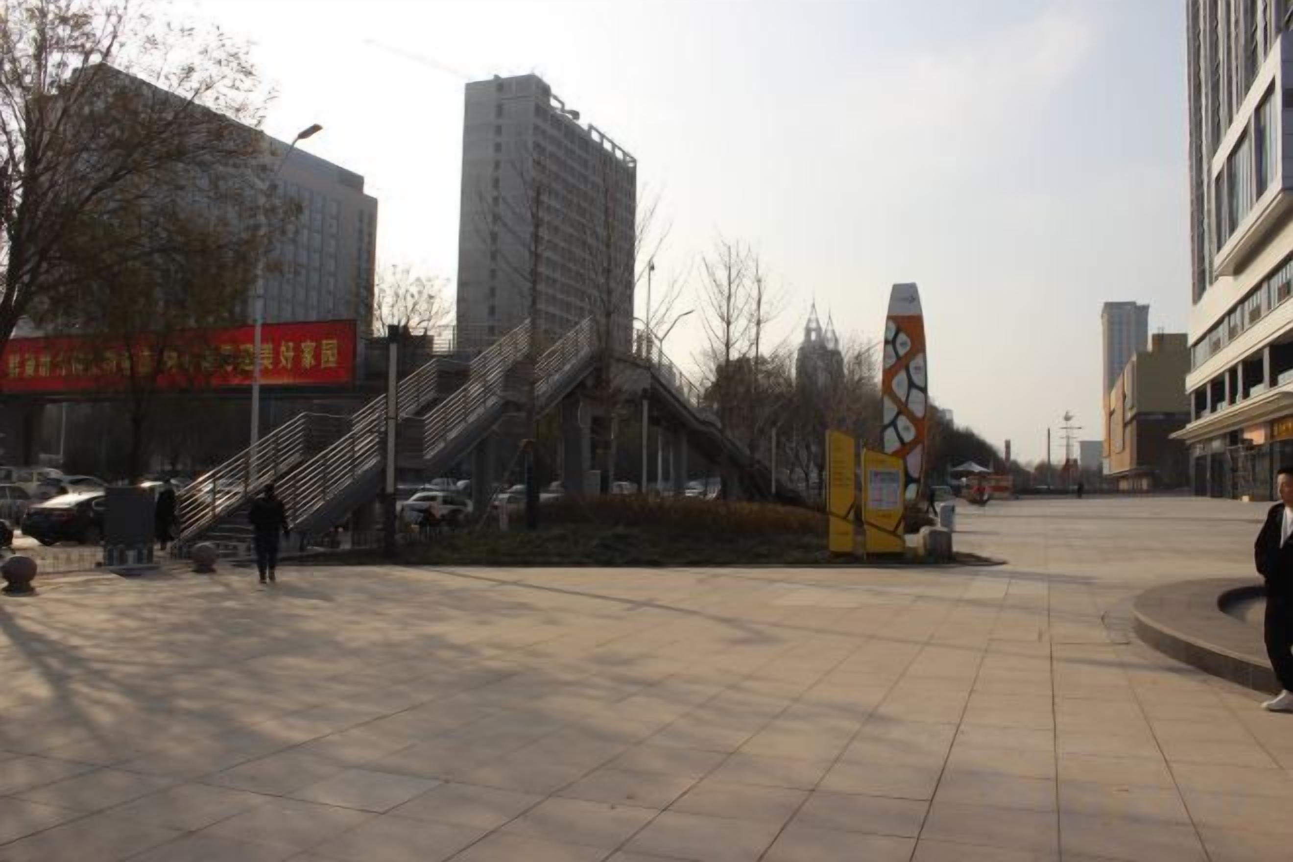 A public square with a staircase leading to a building, a large yellow sculpture, and a red sign.