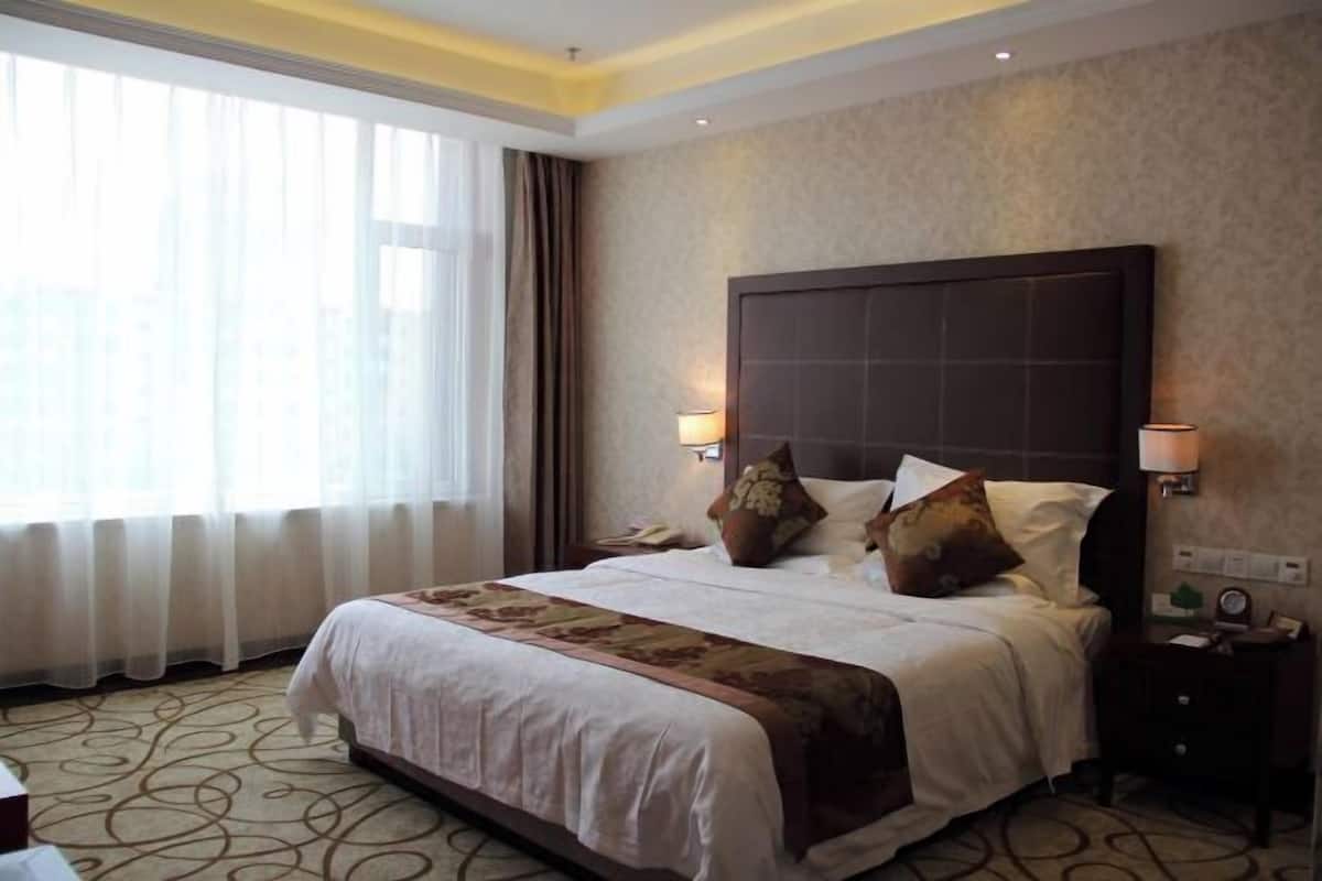 Quarto de hotel com cama grande, duas luminárias de cabeceira, mesa de apoio e janela com cortinas leves.
