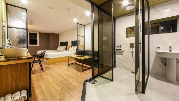 Habitación de hotel moderna con piso de madera, un lavamanos blanco grande, un espejo, una cama con cabecera negra y una pared de vidrio que separa el dormitorio del baño.