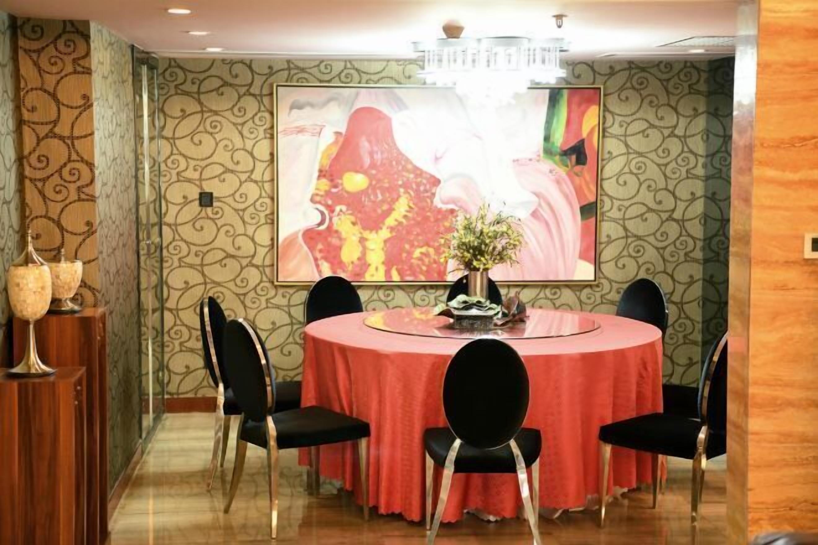 A dining area with a round table covered in a red tablecloth, surrounded by black chairs. There is a chandelier above the table and a large framed artwork on the wall behind it.