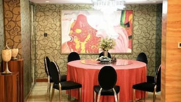 A dining area with a round table covered in a red tablecloth, surrounded by black chairs. There is a chandelier above the table and a large framed artwork on the wall behind it.