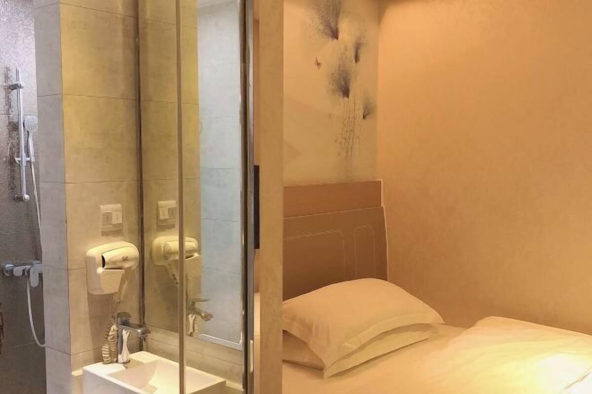 A modern bathroom with a glass shower enclosure, a white sink, and a bed with a beige headboard.