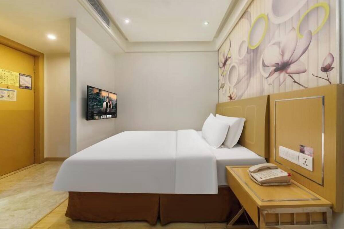 A hotel room with a bed, a bedside table with a phone, a wall-mounted TV, and a floral mural.