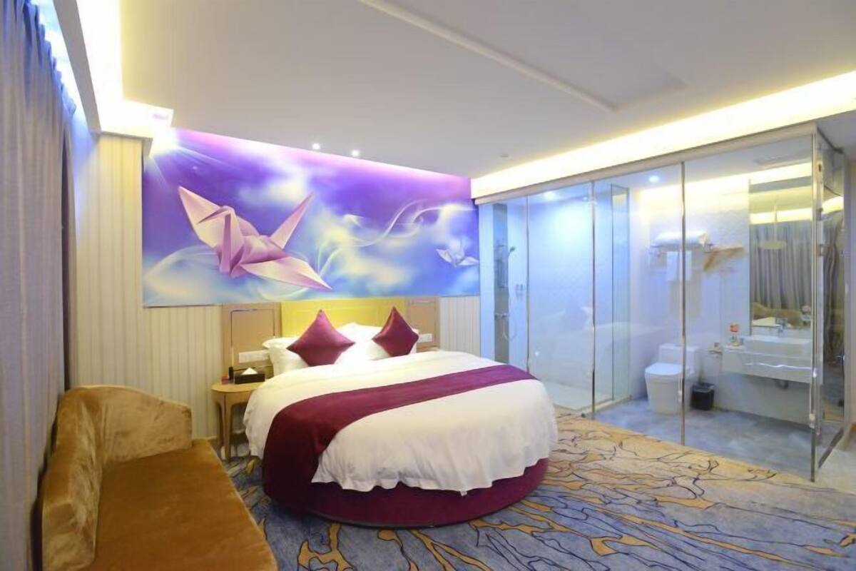 A hotel room with a large bed, a round seating area, a glass-enclosed bathroom, and a wall mural.