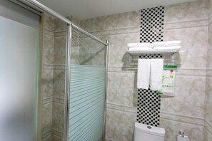 A bathroom with a glass shower enclosure, a toilet, and a shelf with towels and a tissue box. - Sunjiang Hotel (Quanzhou)