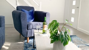 Una sala de estar moderna con un sillón azul, una mesita de centro blanca, una botella de vino y una planta verde grande.