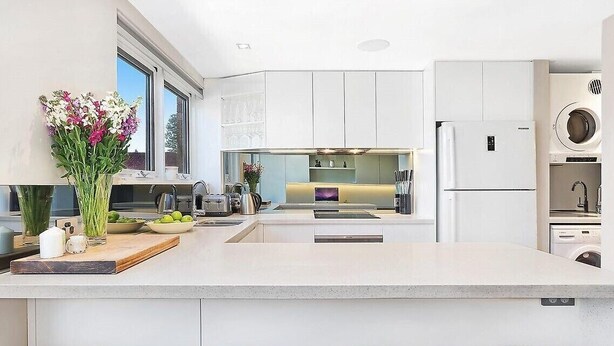 A modern kitchen with white cabinets, a central island with stools, a built-in oven, and a washing machine. - Manly - Free parking, WIFI, Office, Pool, Coffee Machine (Manly)