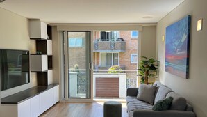 A modern living room with a flat-screen TV, a grey sofa, a round ottoman, and a balcony with a view of a brick building. - Manly - Free parking, WIFI, Office, Pool, Coffee Machine (Manly)