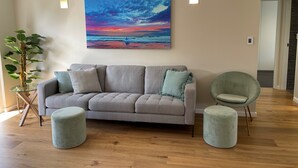 A living room with a grey sofa, two ottomans, a green armchair, a potted plant, and a painting of a sunset. - Manly - Free parking, WIFI, Office, Pool, Coffee Machine (Manly)