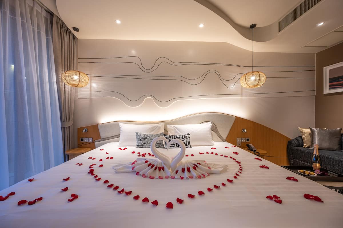 A hotel room with a bed, a bedside table, a nightstand, a sofa, and a wall-mounted light fixture.