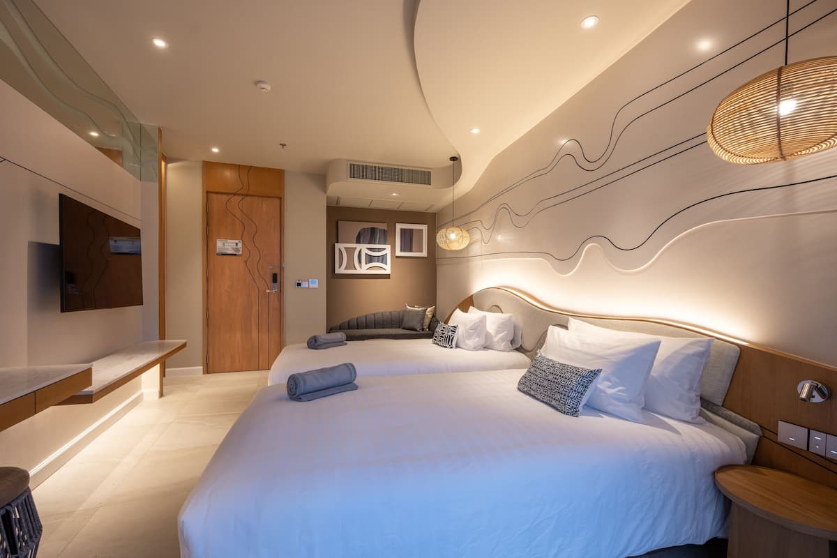 A modern hotel room with a large bed, a TV, a bench, and a wall-mounted light fixture.
