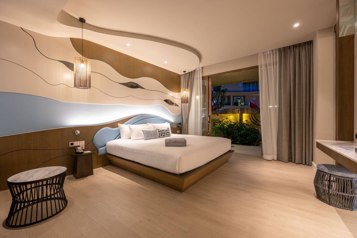 A modern hotel room with a large bed, a bedside table with a phone, a round table, and a view of the outdoors.