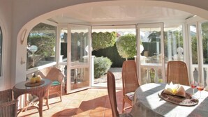 A sunlit patio with a table set for two, surrounded by wicker chairs, and a glass door leading to a garden.