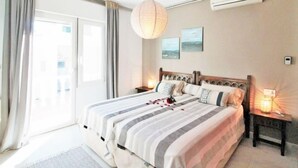 A bedroom with a large bed, a bedside table with a lamp, a window with curtains, and an air conditioning unit on the wall.