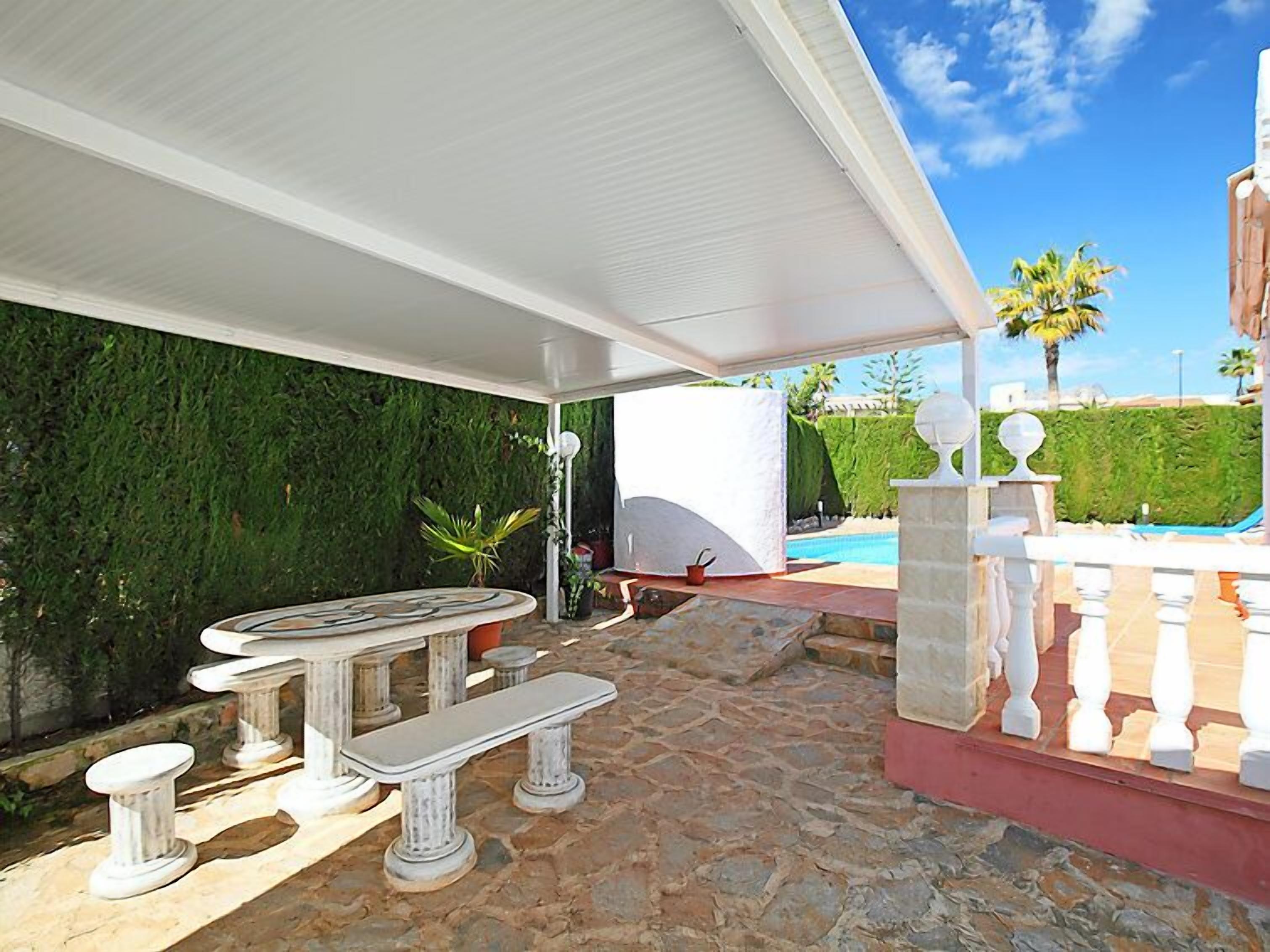 An outdoor patio with a stone floor, a white table with benches, and a view of a swimming pool and greenery.