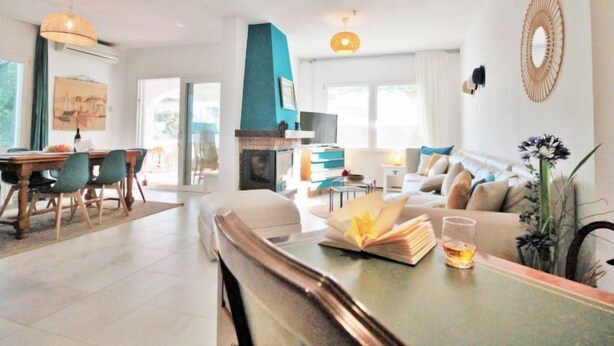 A modern living room with a fireplace, a dining area with a wooden table and chairs, and a sofa with cushions.