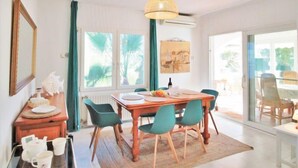 A dining area with a wooden table, green chairs, a hanging light fixture, and a view through large windows.