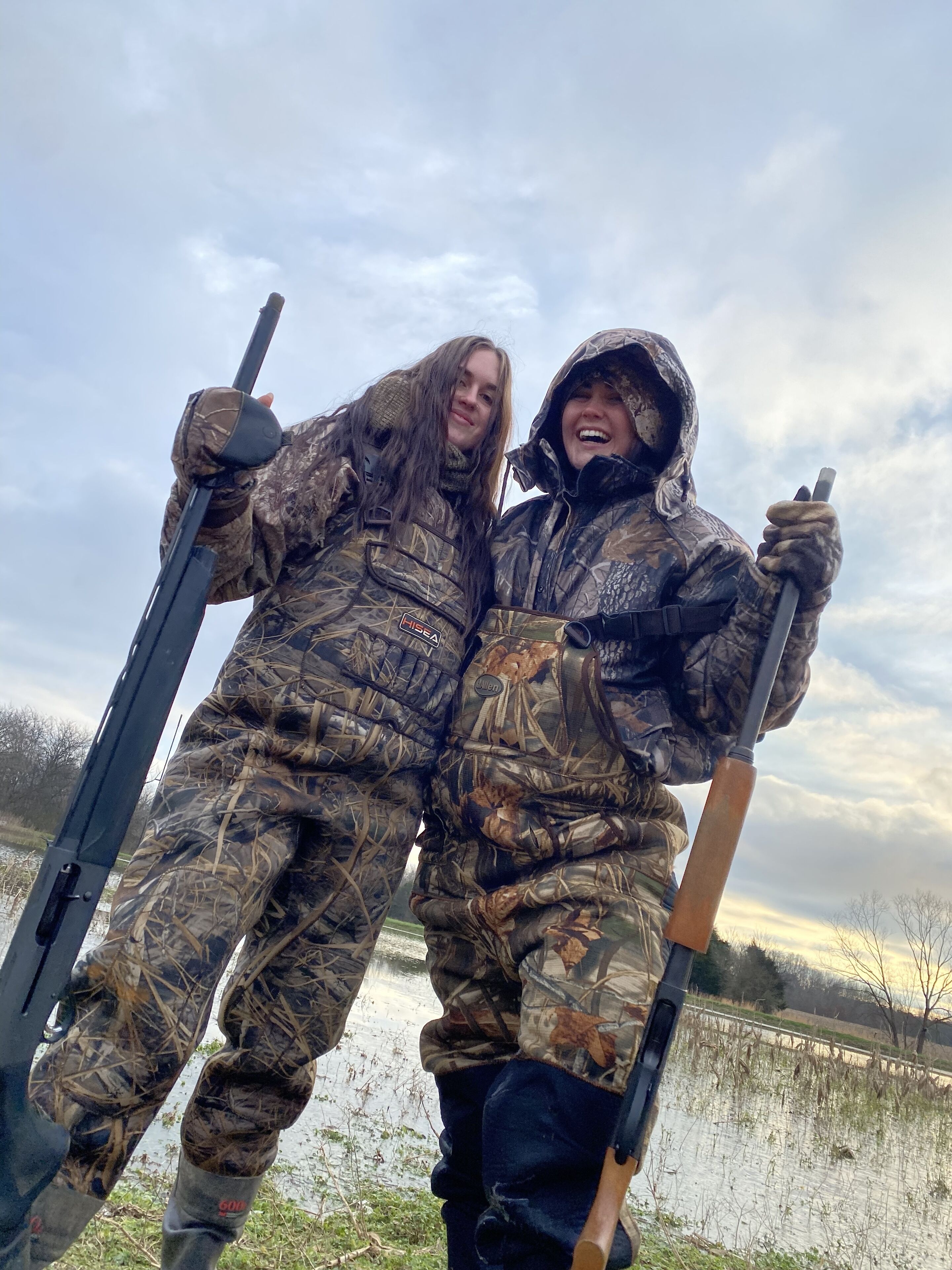 Two people in camouflage clothing holding rifles, standing by a body of water.