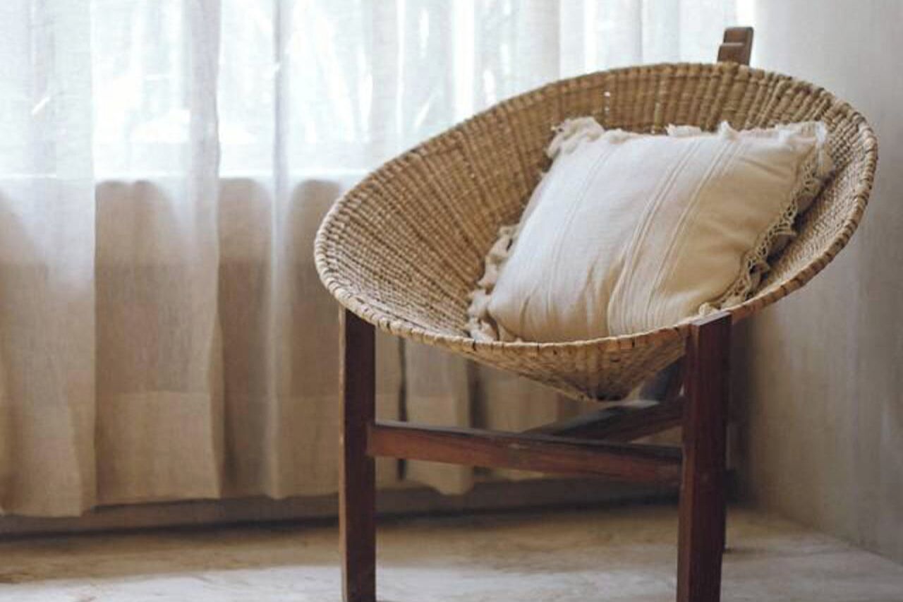 A wicker chair with a cushion, positioned in a room with a radiator and sheer curtains.
