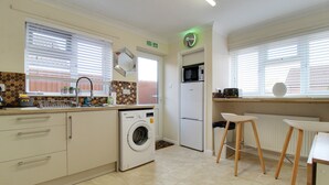 A compact kitchen with a washing machine, a microwave, and a refrigerator. There are two stools and a small table. - Cedar Drive, Holbeach - 1 to 4 - Self Catering (Spalding)