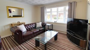 A living room with a leather sofa, a glass coffee table, a flat-screen TV, a large window with curtains, and a ceiling with a decorative pattern. - Cedar Drive, Holbeach - 1 to 4 - Self Catering (Spalding)