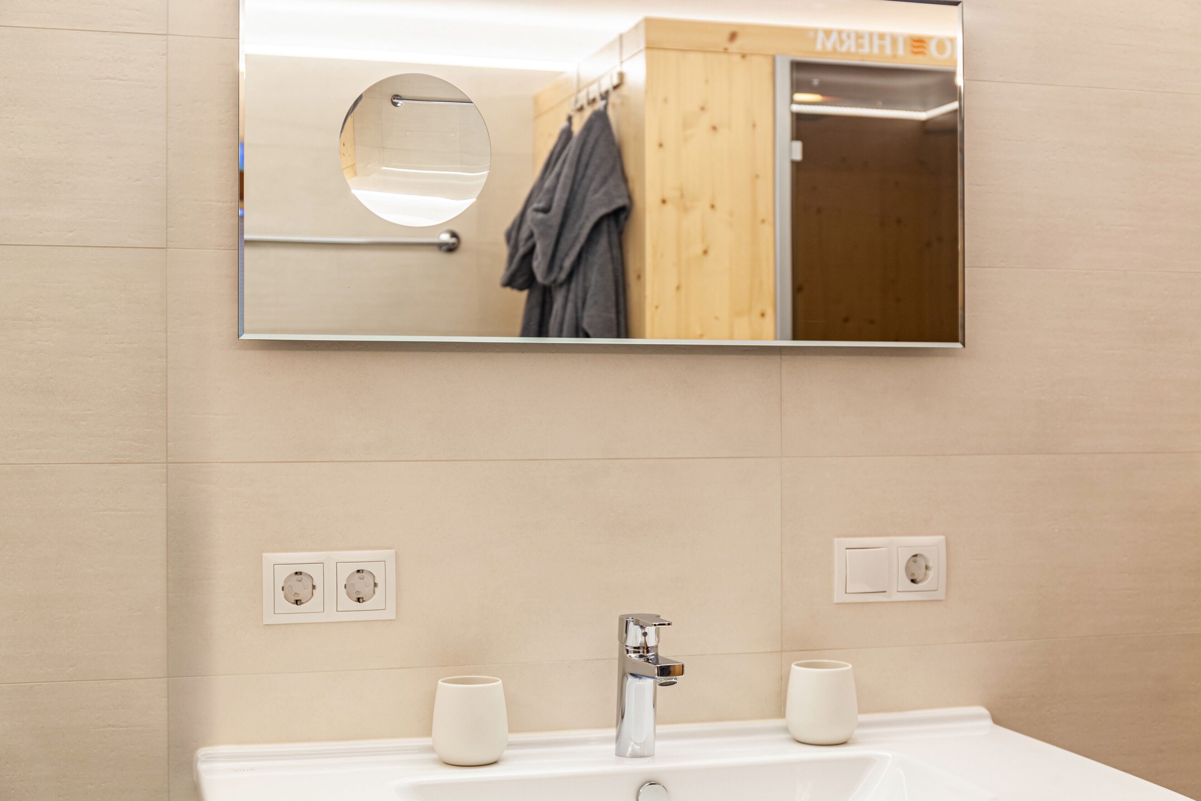 Un baño con un espejo grande, un lavamanos, dos tazas y un toallero.